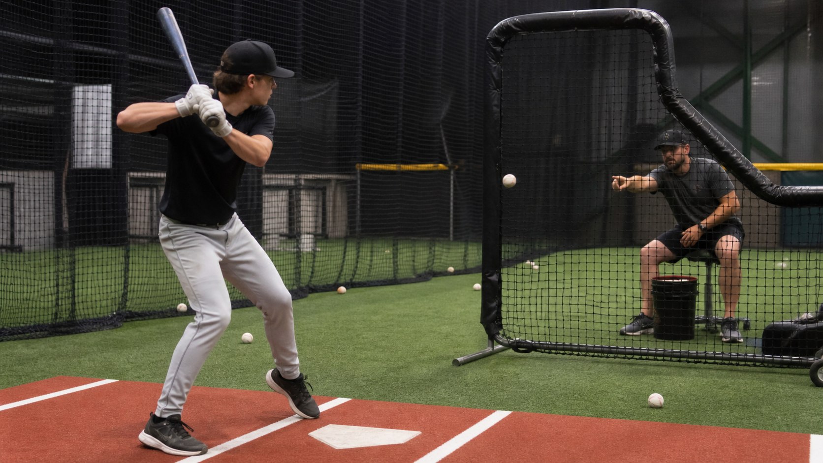 Coach delivering soft toss to a young baseball hitter inside a batting cage during focused mechanics practice