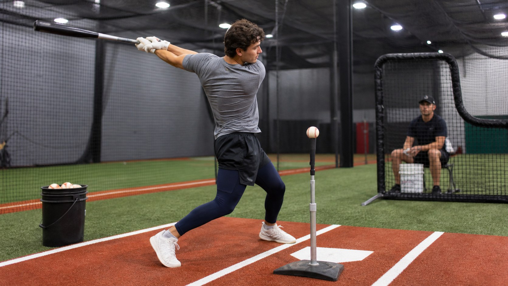 Baseball hitter practicing focused tee work in a batting cage with a coach observing from behind the screen
